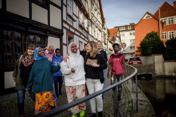eine Gruppe ehrenamtlicher Unterstützerinnen läuft mit vier jungen Geflüchteten durch eine malerische Innenstadt, alle haben fröhliche Gesichter